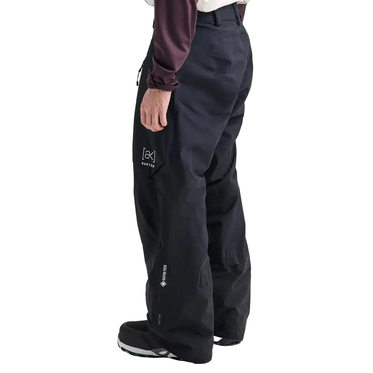 A person wearing black ski pants with a white logo on the left thigh, paired with a dark long-sleeve top and black sneakers, shown from the waist down against a light gray studio background.