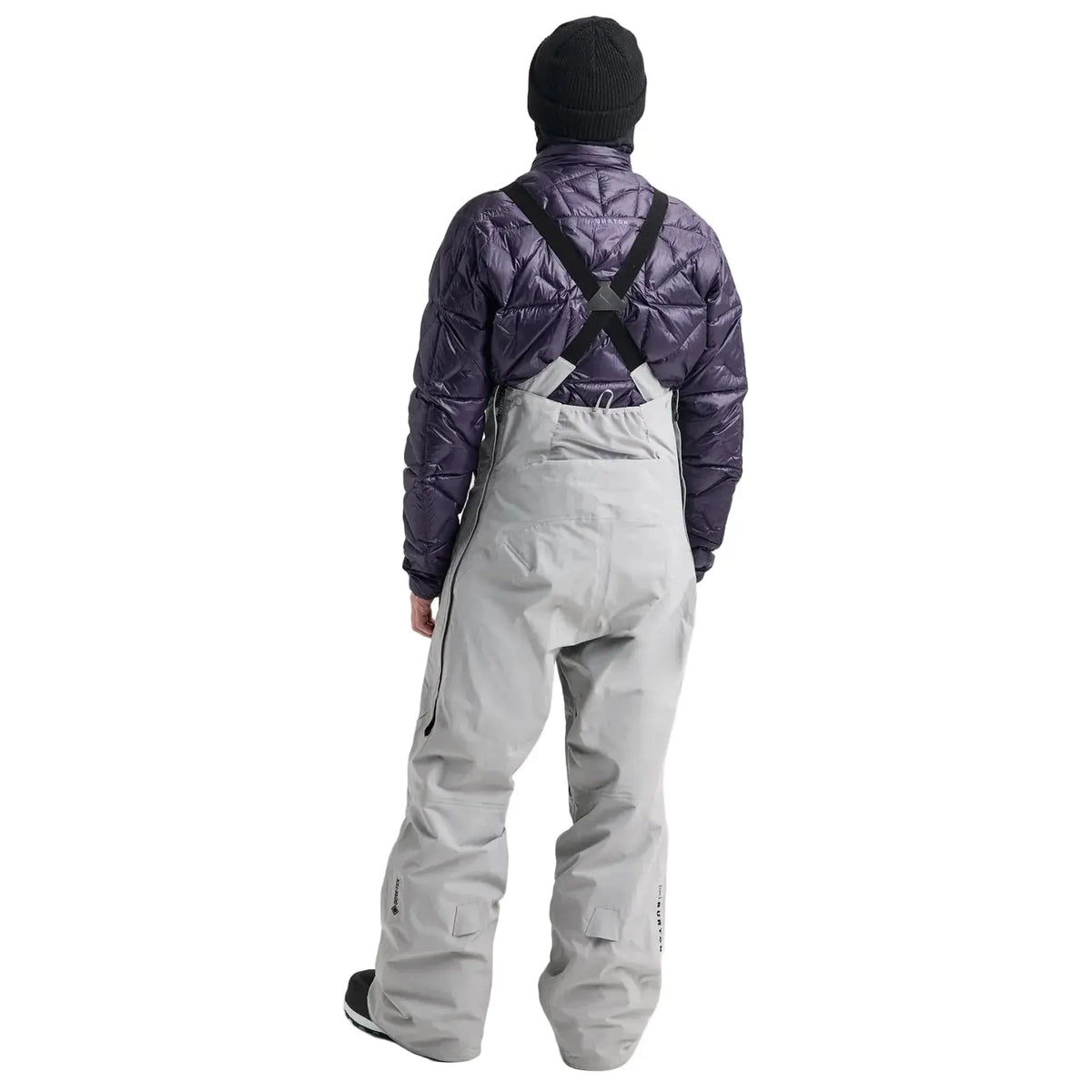 A model viewed from behind wearing a dark purple quilted puffer jacket with black shoulder harness straps and light gray snow pants, paired with a black beanie, on a light gray studio background.