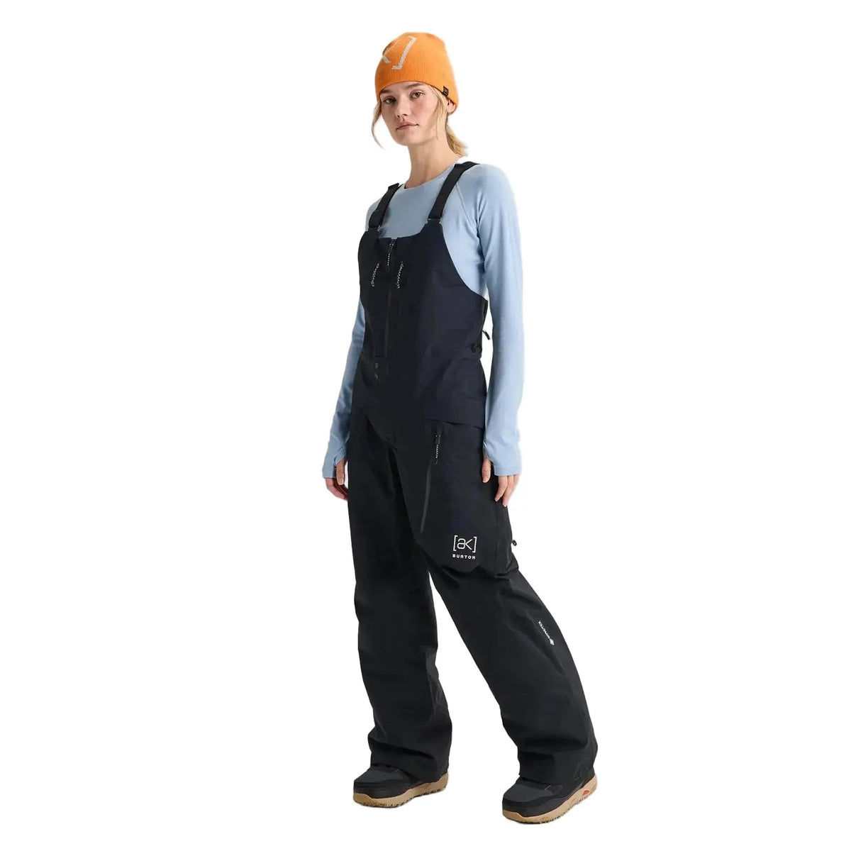 A model wearing black snow bib overalls with a white logo on the thigh, styled over a light blue long-sleeve shirt and an orange beanie, standing against a light gray studio background.