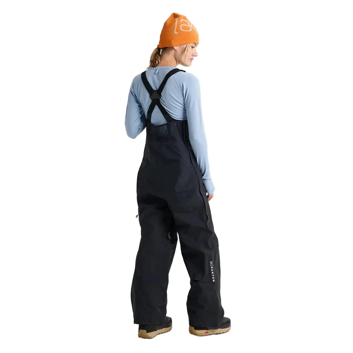 A model shown from the back wearing black snow bib overalls, a light blue long-sleeve shirt, and an orange beanie against a light gray studio background.