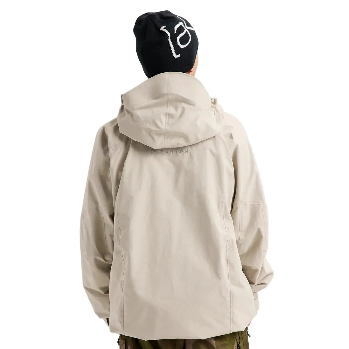 A person shown from the back wearing a light beige hooded jacket and a black beanie with a white graphic, photographed against a plain light gray studio background.