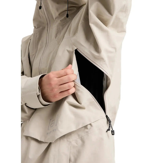 Close-up of a person wearing a light beige technical outdoor jacket, showing a hand opening a side pocket to reveal a black inner layer. The jacket has a subtle logo on the lower pocket flap.