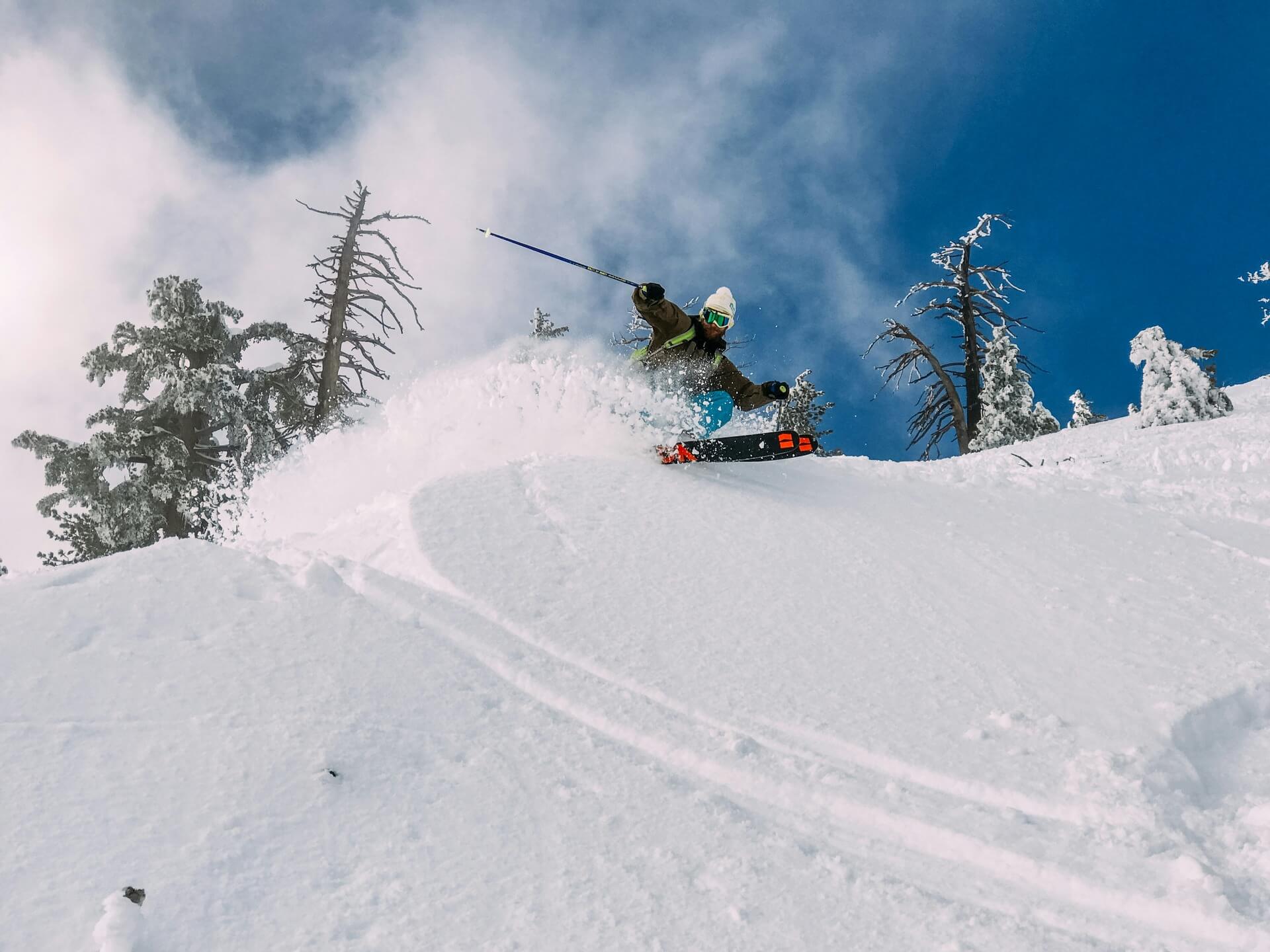 A skier making a sharp turn on a snowy slope with snow spraying up, surrounded by snow-covered trees and a clear blue sky.